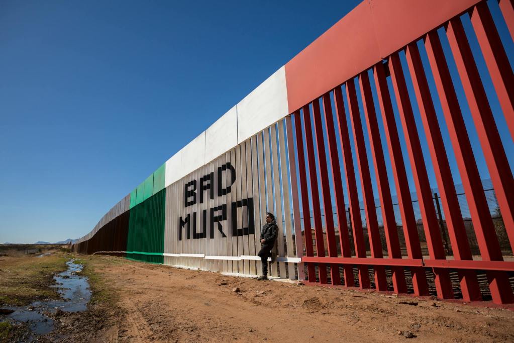 man standing against the wall on the border of usa and mexico with a sign bad muro naco sonora mexico